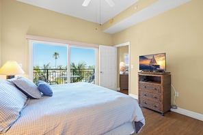Un dormitorio con cama, televisor sobre un mueble, balcón con vistas y puerta que da a otra habitación.