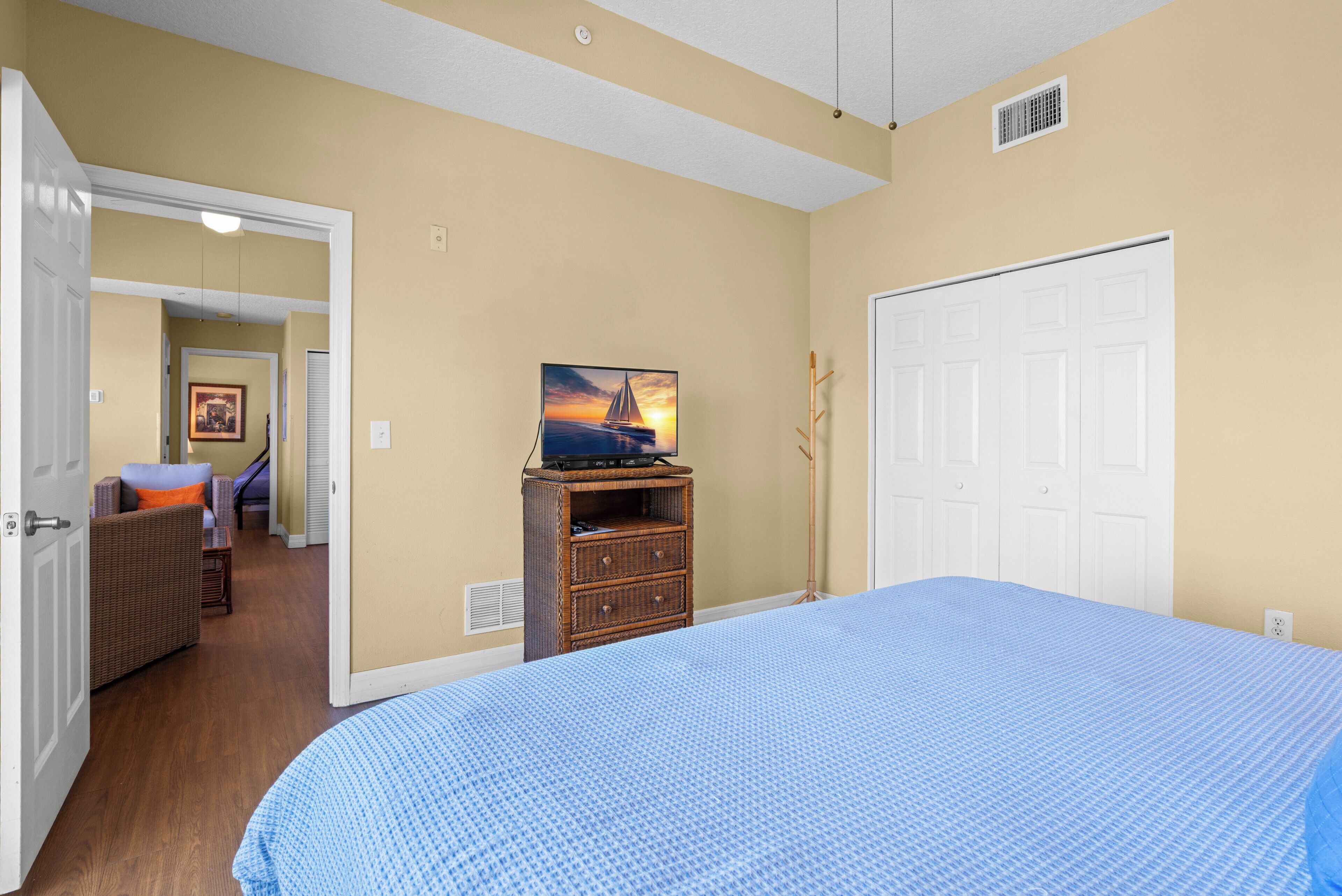 A bedroom with a large bed, a dresser with a television, a door, and a hallway leading to another room.