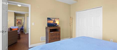 A bedroom with a large bed, a dresser with a television, a door, and a hallway leading to another room.