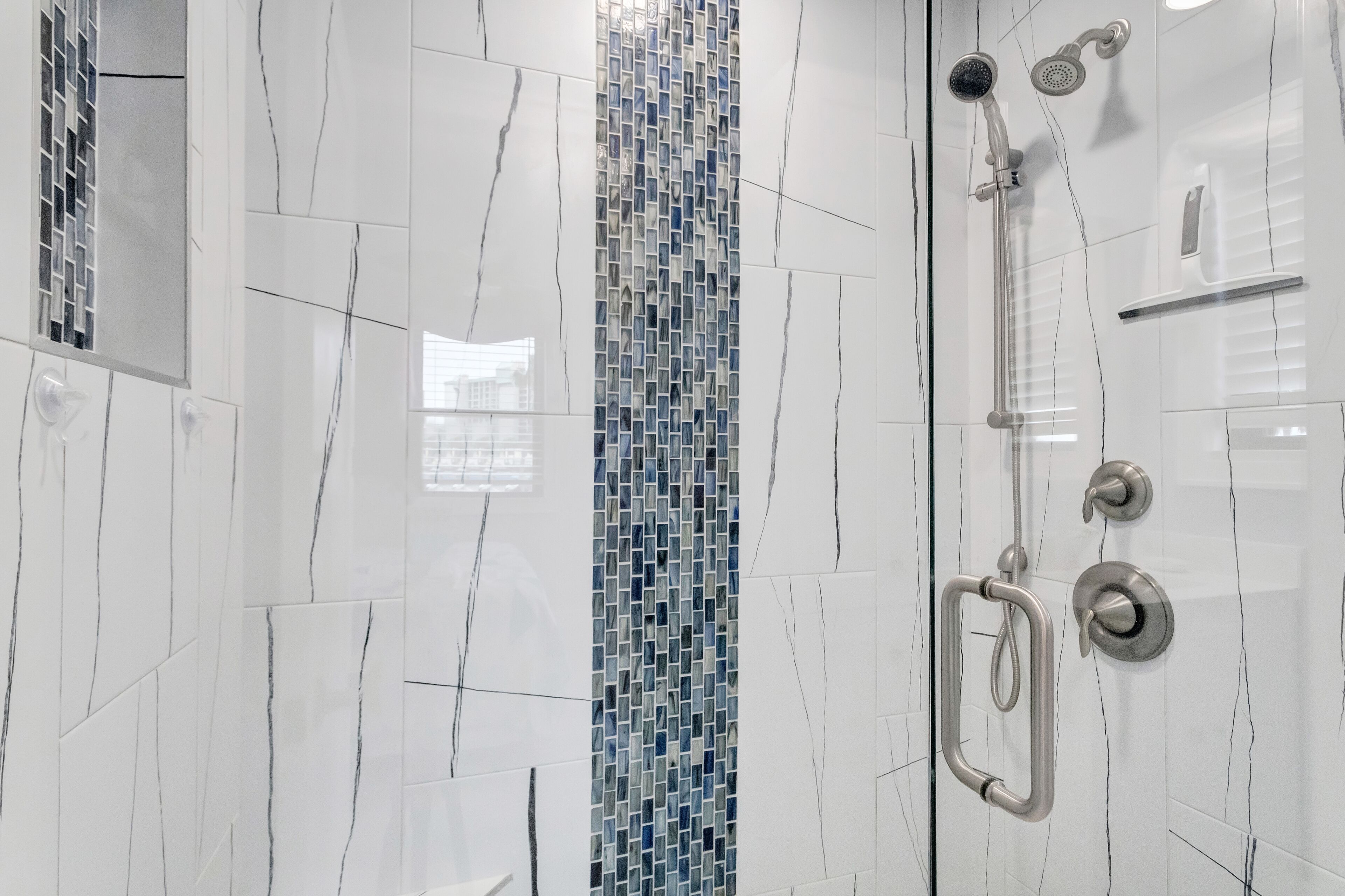 Un baño moderno con baldosas de mármol blanco, un detalle en mosaico azul y una zona de ducha con mampara de cristal.