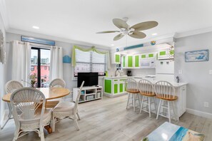 Une cuisine moderne avec des meubles blancs et des touches de vert, un coin repas avec des chaises en osier et une table en bois, et un ventilateur de plafond.