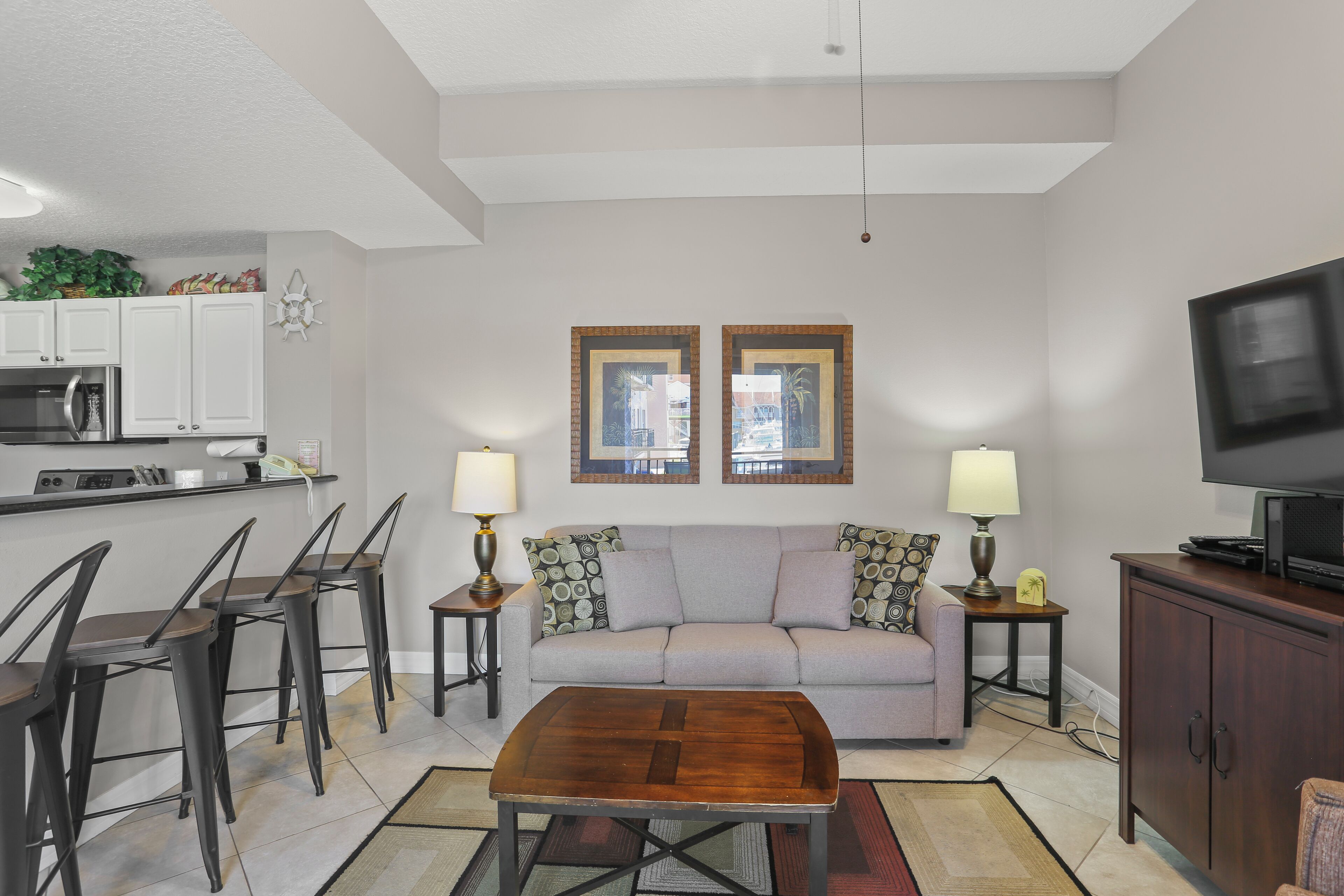 A modern living room with a sofa, coffee table, and TV. There is a kitchen area with cabinets and a microwave.