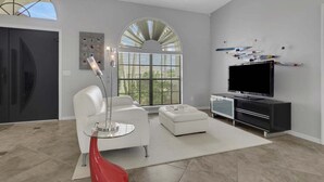 Ein modernes Wohnzimmer mit einem weißen Sofa, einem roten Akzentstuhl, einem Glastisch, einem Flachbildfernseher und einem großen Fenster mit Jalousien.