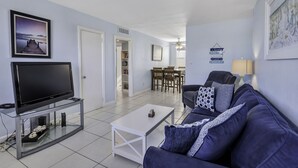 A living room with a flat-screen TV on a glass table, a dark blue sofa with patterned cushions, a white coffee table, and a dining area with a table and chairs. - 🌅 Gulf Winds 2BR Condo — Rooftop Deck with Stunning Beach Views (St Pete Beach)