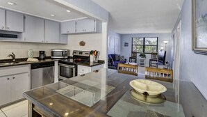 A modern kitchen with stainless steel appliances, a glass dining table, and a view of the living area. - 🌅 Gulf Winds 2BR Condo — Rooftop Deck with Stunning Beach Views (St Pete Beach)