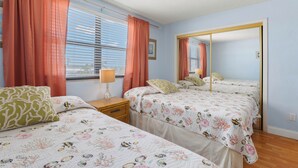 A bedroom with two beds, a window with blinds, a mirror, and a nightstand with a lamp. - Gulf Winds 1505 — Updated Tile Floors & Room for Everyone (St Pete Beach)