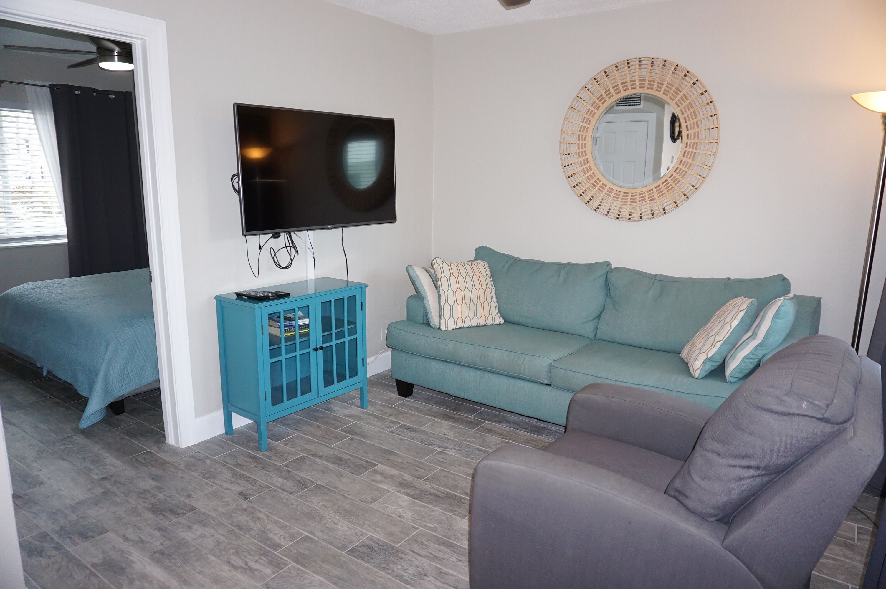A living room with a teal sofa, a grey armchair, a TV, a small blue cabinet, and a round mirror on the wall.