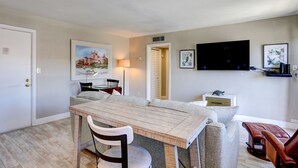 A dining area with a wooden table and chairs, a sofa, and a television mounted on the wall. - 🌅 Gulf Winds 2nd-Floor Condo — Gulf Views & Bright Beachside Charm (ST PETE BEACH)