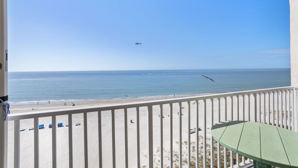 A balcony with a view of the beach and ocean, a green table, and a white railing.