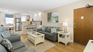 A modern living room with a grey sofa, a coffee table, and a TV. There is a kitchen in the background with white cabinets and a refrigerator. - 🌊 Gulf Winds 2BR Condo — Partial Gulf Views, Remodeled Bath & Steps to the Sand (ST PETE BEACH)