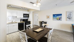 Une cuisine moderne avec des armoires blanches, une cuisinière noire et un coin repas comprenant une table en bois foncé et des chaises blanches. Des luminaires sont fixés au plafond et des œuvres d’art ornent les murs.