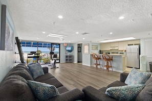 A modern living room with a sofa, a dining area with bar stools, and a kitchen with a refrigerator and microwave. - 🌊 Caprice Resort Unit #302 – Gulf Front Comfort with Room for Six (St Pete Beach)
