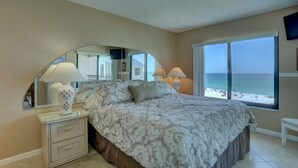 A bedroom with a large bed, a nightstand with lamps, a mirror, and a view of the beach. - 🌞 Caprice Resort Unit #502 – Renovated Gulf Front Escape for Four Guests (St Pete Beach)