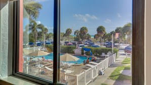 On peut voir une piscine entourée de chaises longues et de parasols, un parking avec des voitures, et des palmiers.