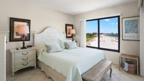 A bedroom with a large window offering a beach view, a bed with a light-colored quilt, bedside tables with lamps, and a small bench.