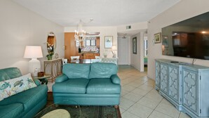 A living room with a teal sofa, a television, and a kitchen in the background.