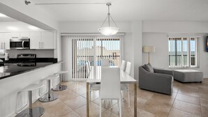 A modern kitchen with a dining area, a gray sofa, and a large window with blinds. - 🌅 Villa Madeira #601 — Corner Unit with Wraparound Balcony — Gulf & Bay Views (Madeira Beach)