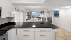 A modern kitchen with white cabinetry, a black countertop, and a dining area with a white table and chairs. - 🌅 Villa Madeira #601 — Corner Unit with Wraparound Balcony — Gulf & Bay Views (Madeira Beach)