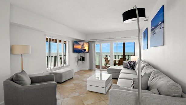 A modern living room with a sofa, a coffee table, and a TV. There are large windows with a view of the sea. - 🌅 Villa Madeira #601 — Corner Unit with Wraparound Balcony — Gulf & Bay Views (Madeira Beach)