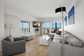 A modern living room with a sofa, a coffee table, and a TV. There are large windows with a view of the sea.