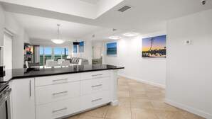 A modern kitchen with white cabinetry, black countertops, and a large island. There are multiple windows providing natural light and a view of the outdoors. - 🌅 Villa Madeira #601 — Corner Unit with Wraparound Balcony — Gulf & Bay Views (Madeira Beach)