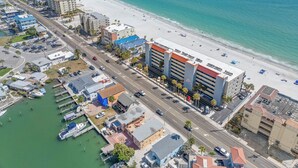 An aerial view of a coastal city with a marina, a beach, and a hotel with multiple floors and balconies. - 🌅 Villa Madeira #601 — Corner Unit with Wraparound Balcony — Gulf & Bay Views (Madeira Beach)