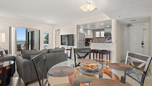 A modern living room with a glass dining table, grey sofa, and a kitchen in the background.