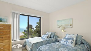 A bedroom with two beds, a window with a view of the sea, a wooden dresser, and a lamp.