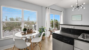 Una cocina moderna con lavavajillas, un comedor con mesa redonda y sillas blancas, y ventanas grandes con persianas.