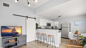 Una cocina moderna con armarios blancos, un microondas y un refrigerador. Hay tres taburetes de bar y una mesa de comedor con sillas. Un televisor está sobre un mueble en la zona de estar.