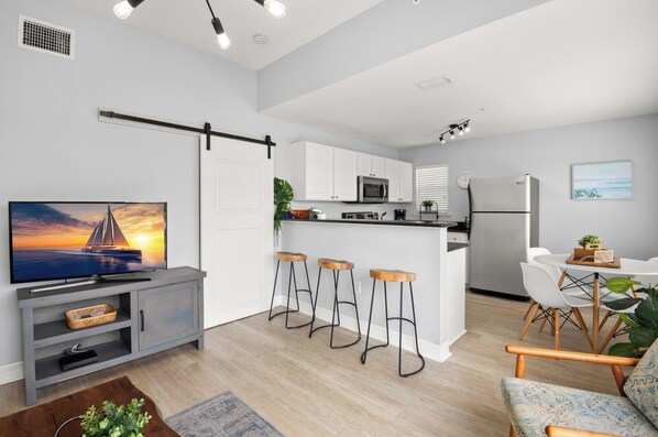 Una cocina moderna con armarios blancos, un microondas y un refrigerador. Hay tres taburetes de bar y una mesa de comedor con sillas. Un televisor está sobre un mueble en la zona de estar.
