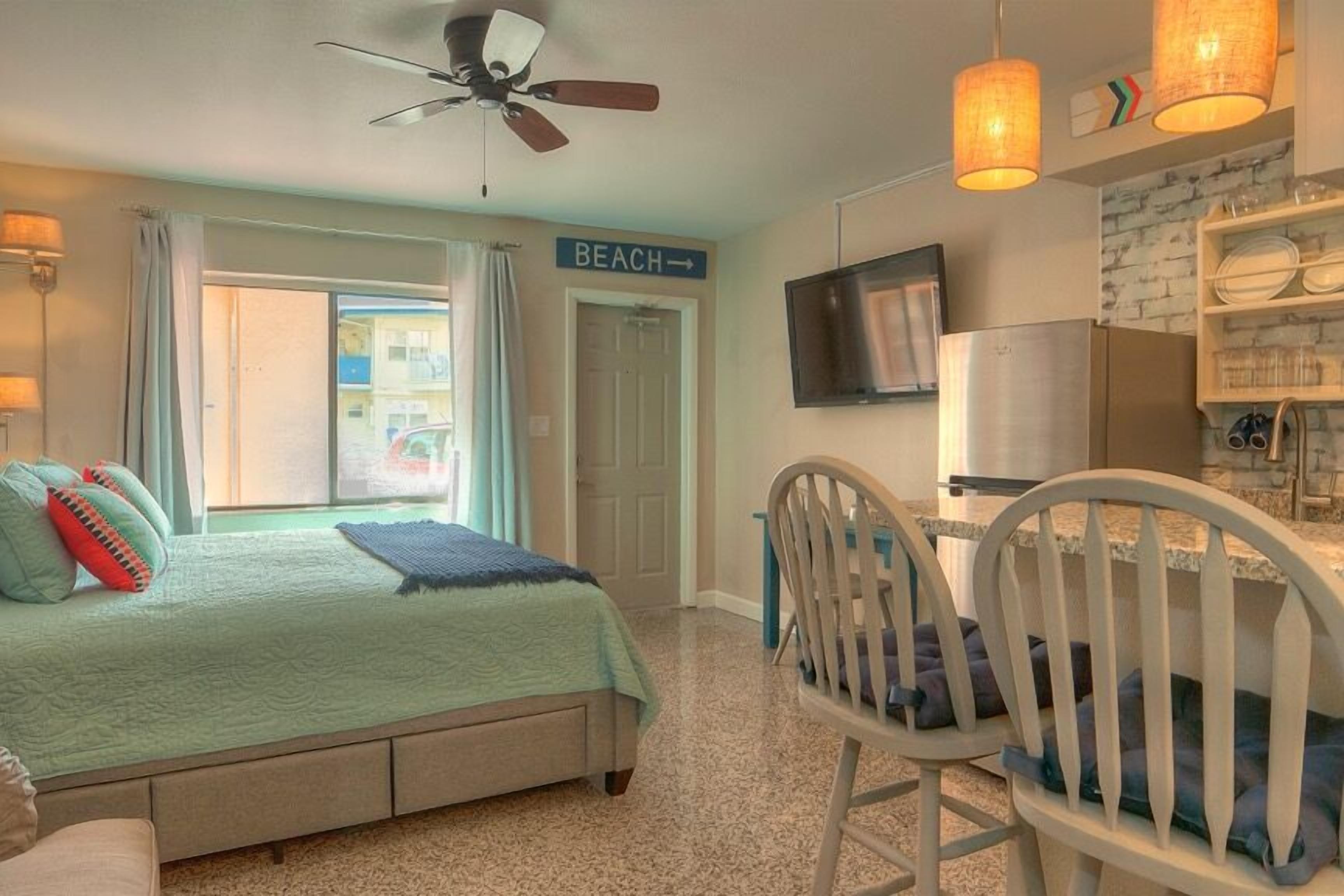 Un dormitorio con cama, escritorio, silla, ventilador de techo, televisor y un cartel que indica la dirección a la playa.