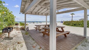A beachside wooden pavilion with picnic tables, surrounded by sand and vegetation, overlooking the beach.