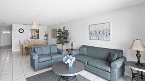 Un salon avec un ensemble de canapés gris, une table basse ronde ornée de fleurs, un coin repas avec des chaises en bois et un tableau représentant un paysage urbain accroché au mur.