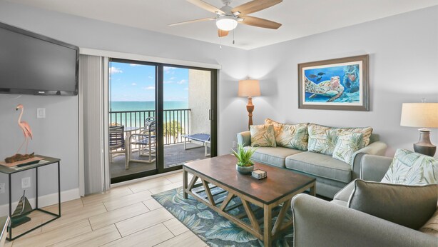 A living room with a ceiling fan, a flat-screen TV, a sofa with patterned cushions, a coffee table with a plant, and a view of the ocean through a sliding glass door.