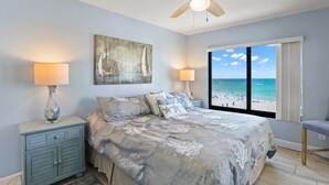 A bedroom with a bed, two bedside lamps, a ceiling fan, a nightstand, a beach view, and a chair.