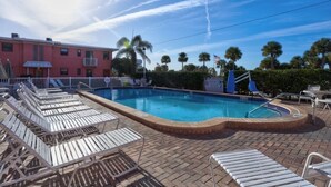 A swimming pool with lounge chairs, a brick patio, palm trees, and a building in the background.