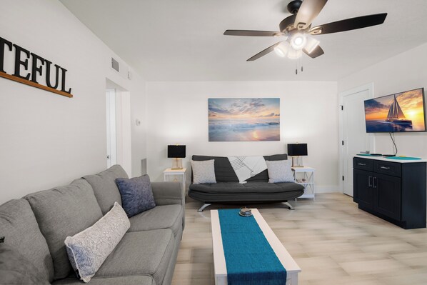 A modern living room with a grey sofa, a black sofa, a coffee table, a ceiling fan, a TV, and artwork on the wall. - 🌿 Gulf Winds 1BR Garden Condo — Tiled Comfort & Flexible Sleeping Options (St Pete Beach)