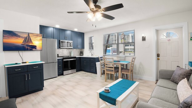 A modern kitchen with dark cabinets, a dining area with a table and chairs, a ceiling fan, and a large window with blinds.