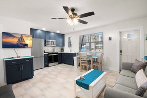 A modern kitchen with dark cabinets, a dining area with a table and chairs, a ceiling fan, and a large window with blinds. - 🌿 Gulf Winds 1BR Garden Condo — Tiled Comfort & Flexible Sleeping Options (St Pete Beach)
