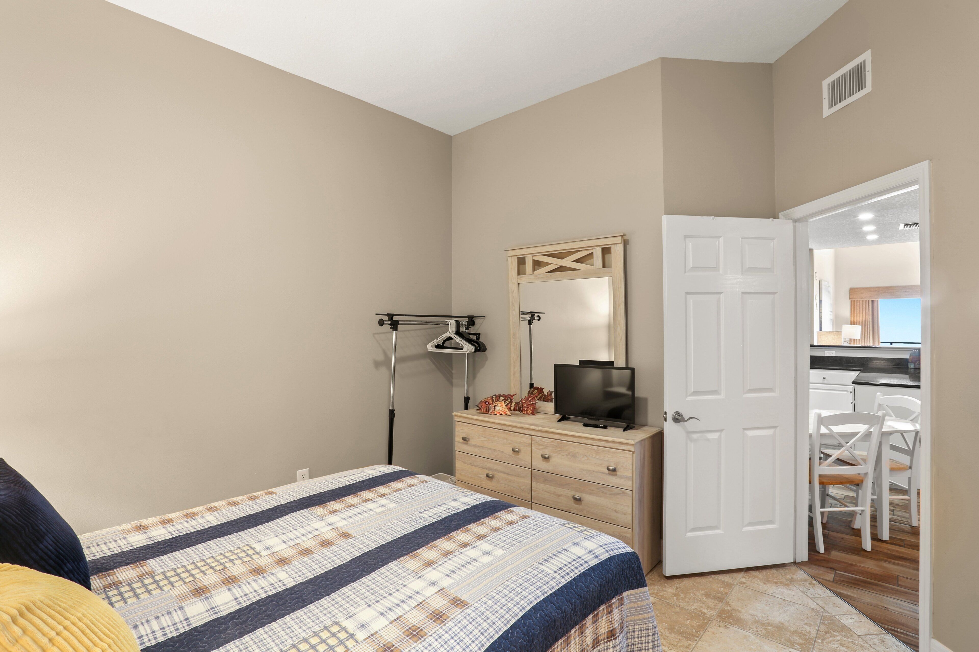 A bedroom with a bed, dresser, television, and a door leading to a kitchen.