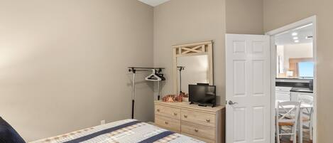 A bedroom with a bed, dresser, television, and a door leading to a kitchen.
