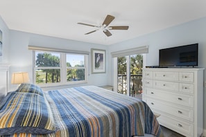 Ein Schlafzimmer mit Bett, Deckenventilator, Kommode, Fernseher und großen Fenstern.