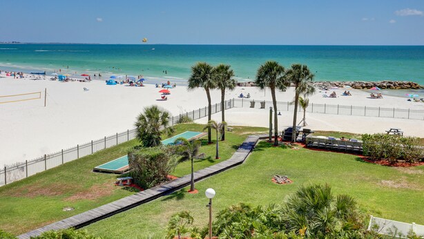 A beach view with a fenced area, palm trees, and a pool. - 🌅 Caprice Resort Unit #303 – Gulf Front Escape for Up to Four Guests (St Pete Beach)