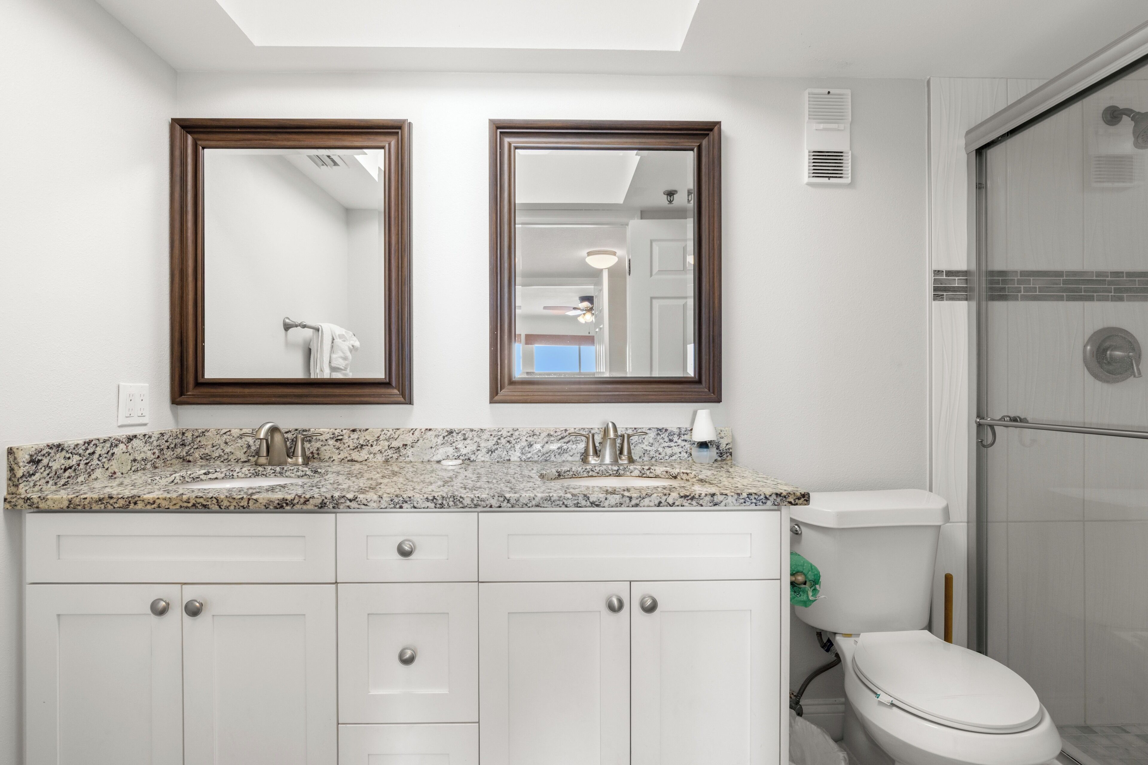 A bathroom with a granite countertop, two sinks, a toilet, and a shower.