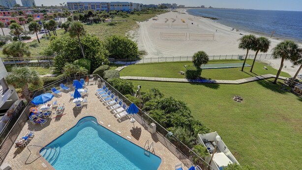Uma área de piscina com espreguiçadeiras e guarda-sóis, uma praia com quadras de voleibol e vista para o oceano.