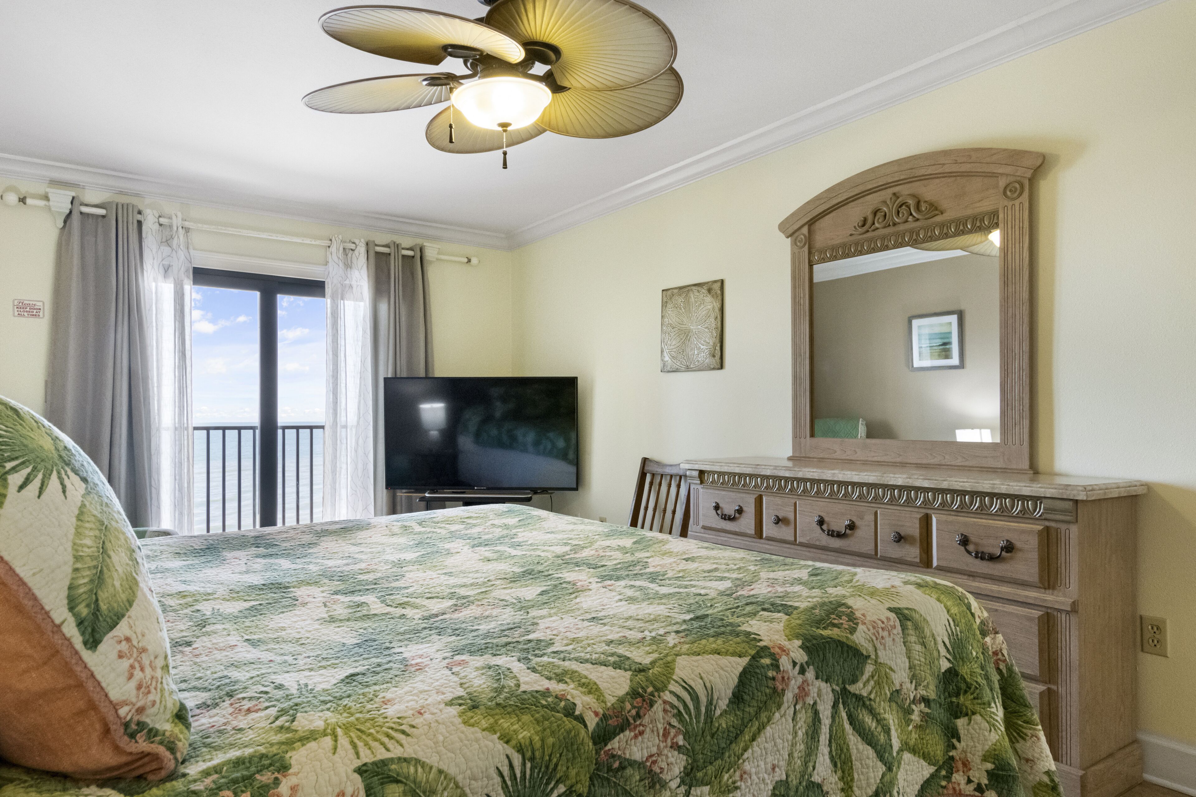 Un dormitorio con cama, cómoda, televisor, ventilador de techo y balcón con cortinas.