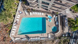 An aerial view of a swimming pool with lounge chairs and a sun deck, surrounded by palm trees and a multi-level building. - 🌅 Prime View Condo — Gulf Strand Resort, St. Pete Beach (St Pete Beach)