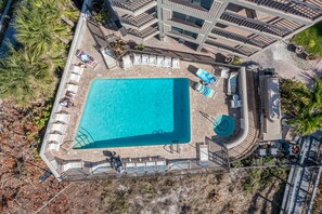 An aerial view of a swimming pool with lounge chairs and a sun deck, surrounded by palm trees and a multi-level building.