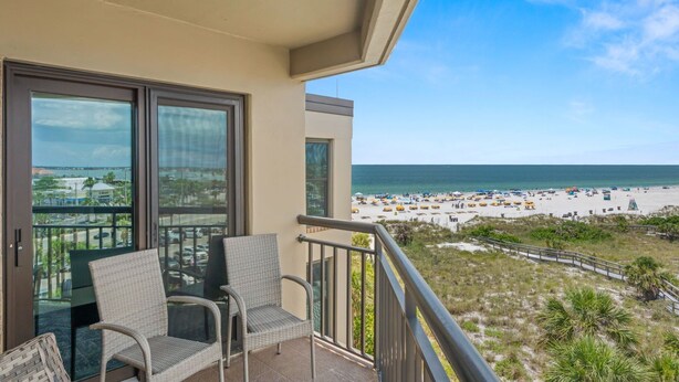 A balcony with wicker chairs, a glass door, and a view of a beach with umbrellas and people. - 🌅 Prime View Condo — Gulf Strand Resort, St. Pete Beach (St Pete Beach)